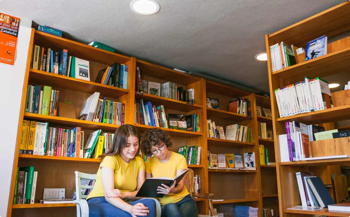 Two young readers share a book in front of tall, full bookshelves.